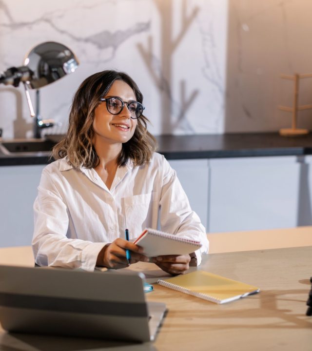Young woman in glasses working indoors with laptop and camera, smiling while taking notes.